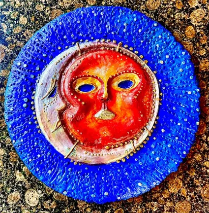 A textured red sun face tucked against a pale crescent moon, inside a ring of thick blue paint on a gold-flecked ground.